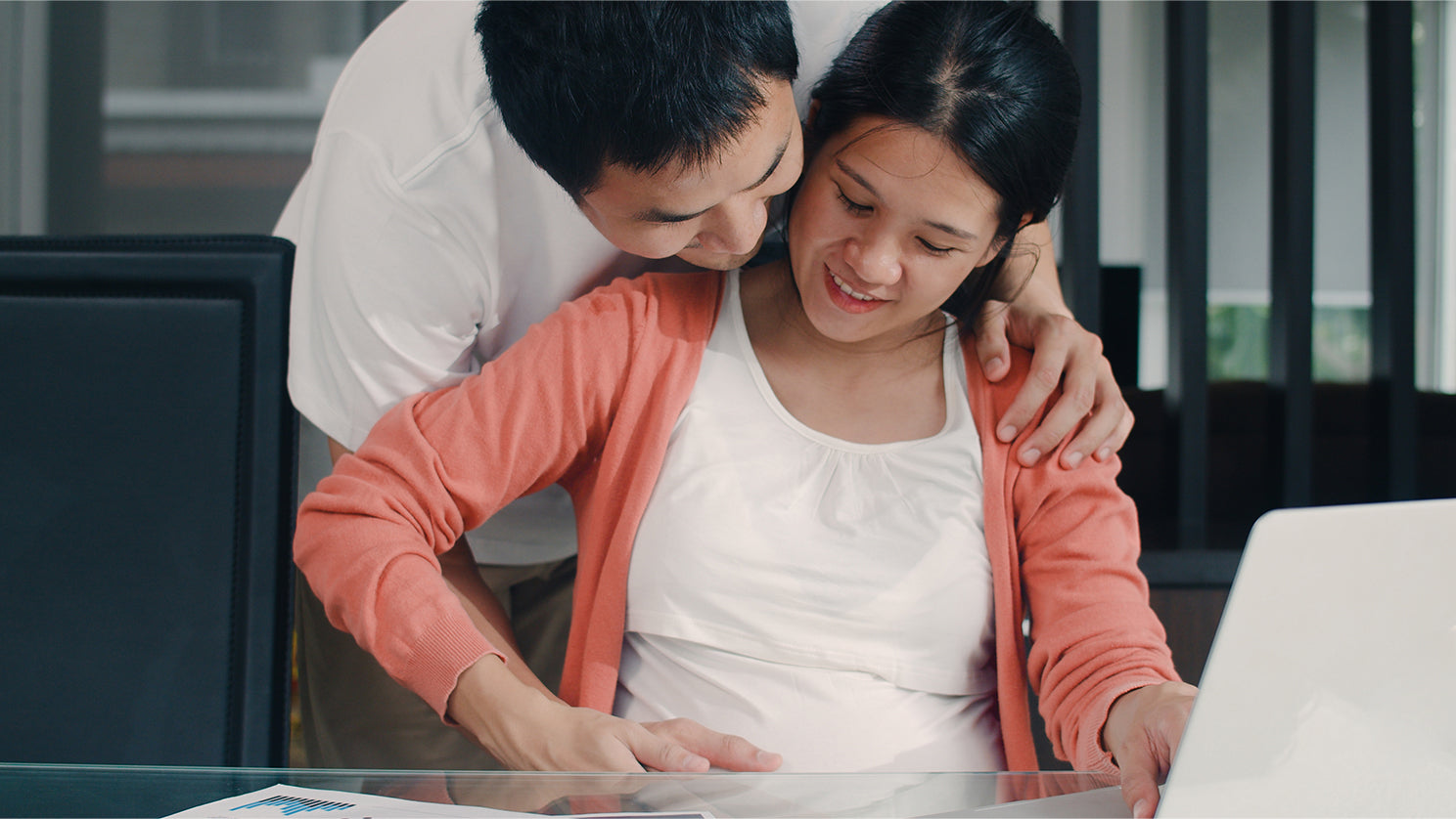 happy young asian pregnant woman using laptop to work and find out about paternity and maternity leave while dad hugs his wife belly