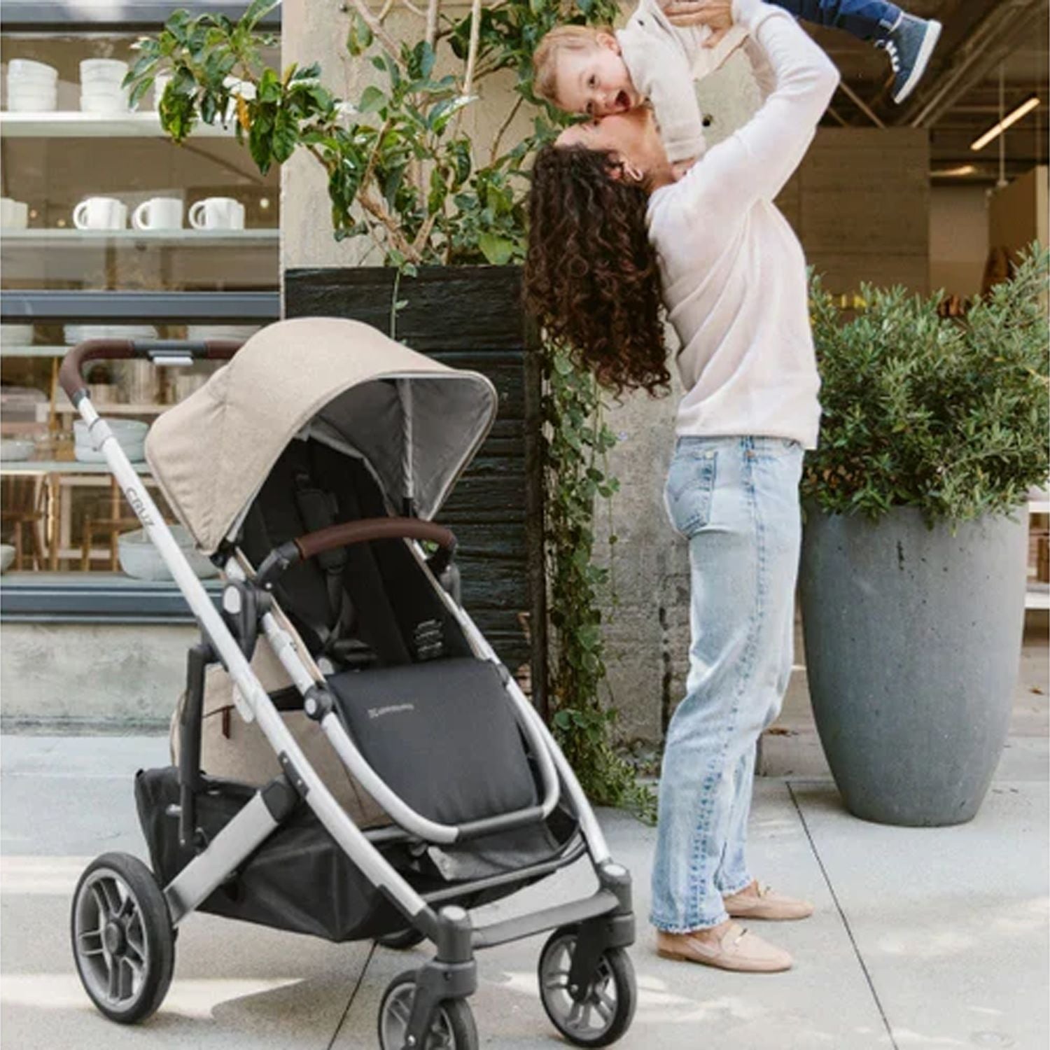 Mother carrying her baby with the Uppababy Cruz 2 Declan Reversible Stroller parked beside